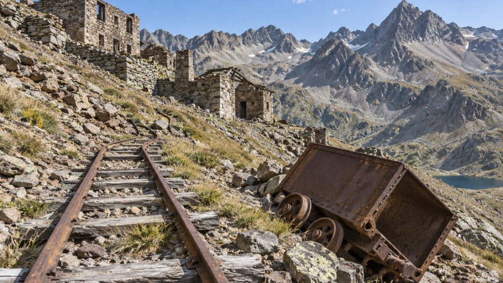Rails de mine à voie étroite rouillés émergeant des éboulis, wagonnet métallique rouillé renversé à côté, bâtiments miniers en ruine sur la pente