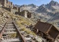 Rails de mine à voie étroite rouillés émergeant des éboulis, wagonnet métallique rouillé renversé à côté, bâtiments miniers en ruine sur la pente