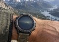 Montre de sport robuste portée au poignet, tenue devant une vallée de montagne floue, poignet et bracelet usés au premier plan