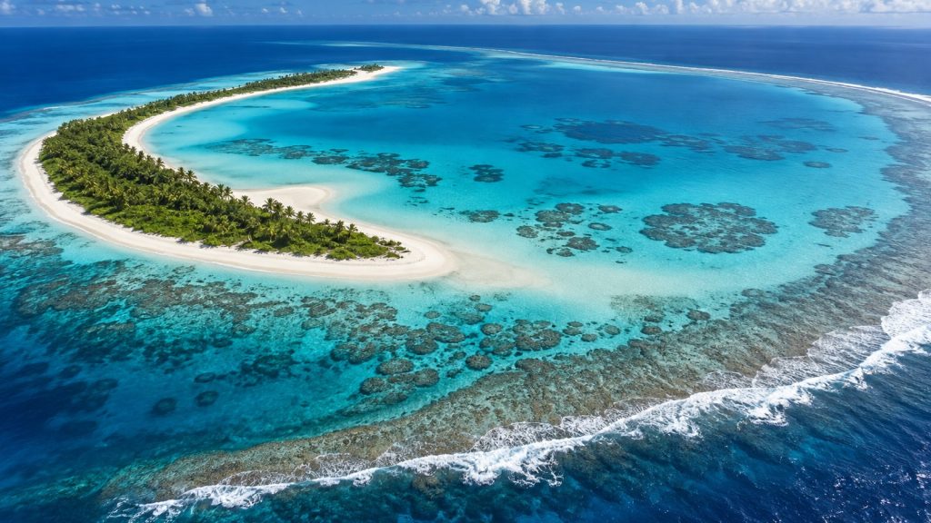 Vue aérienne d'un atoll corallien : croissant de sable blanc et de végétation entourant un lagon turquoise, océan bleu profond au-delà