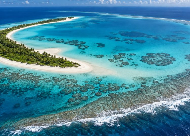 Vue aérienne d'un atoll corallien : croissant de sable blanc et de végétation entourant un lagon turquoise, océan bleu profond au-delà