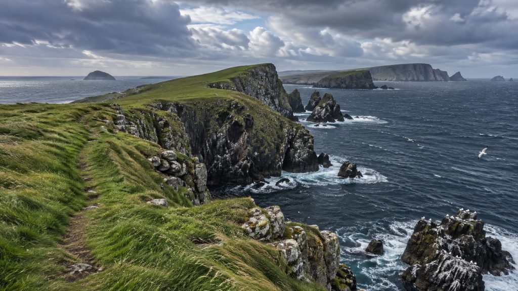 Presqu'île écossaise herbeuse à falaises rocheuses plongeant dans une mer bleu-gris, sentier usé dans l'herbe, ciel de nuages bas